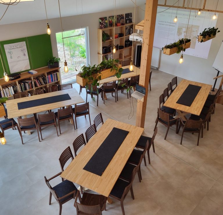 Großer, heller Besprechungsraum mit Tischen und Stühlen, Pflanzen und Fensterlicht.