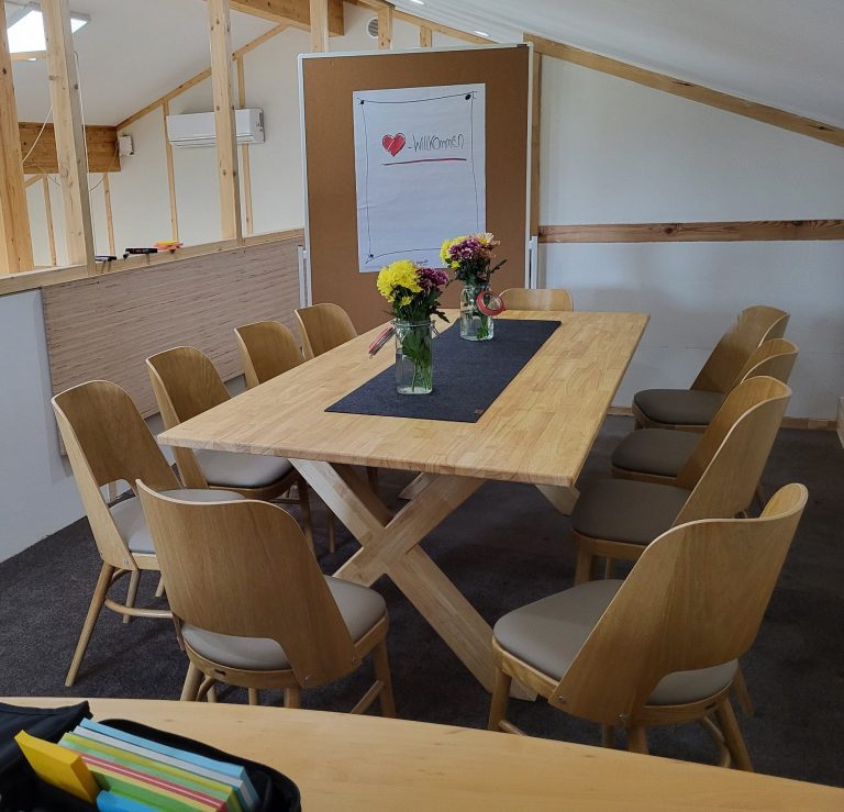 Holztisch umgeben von Stühlen, Blumenvasen und einem Plakat im Hintergrund.