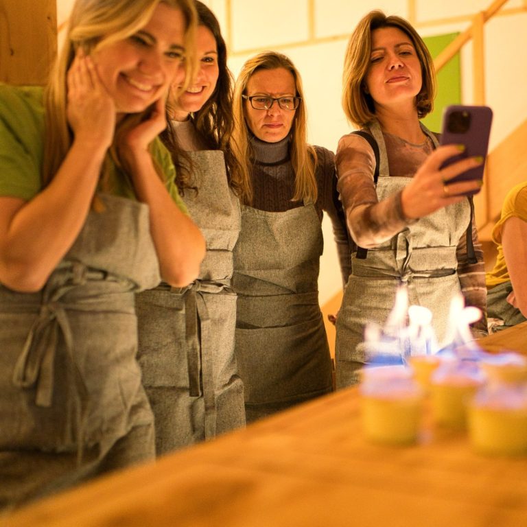 Fünf Frauen in Schürzen lächeln, während sie eine Flamme beobachten.