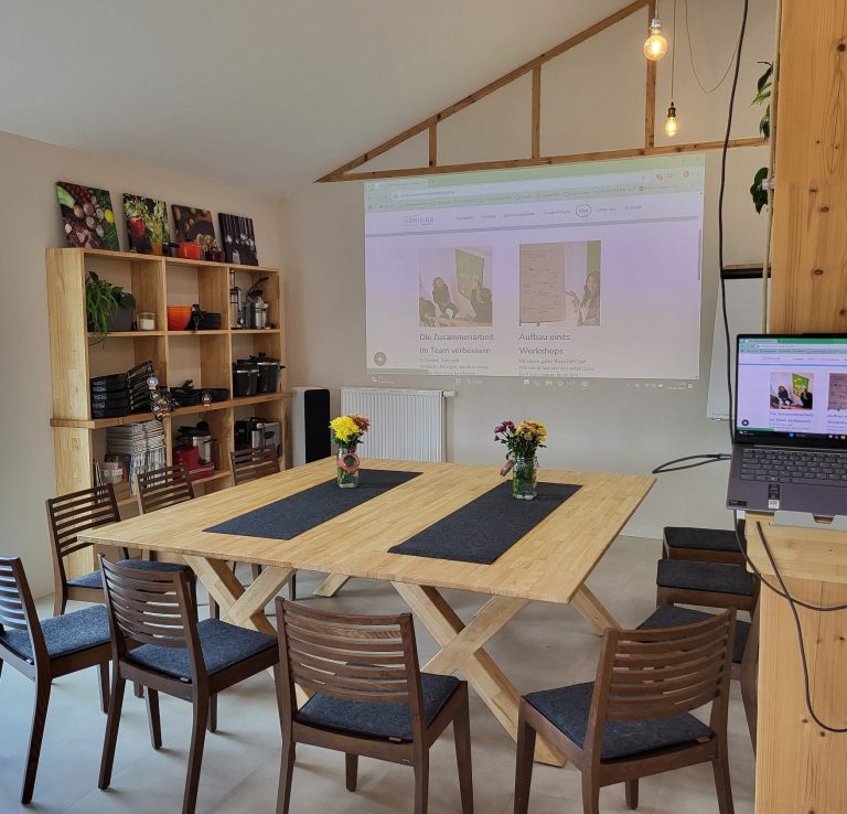 Besprechungsraum mit einem Holztisch, 10 Stühlen, Blumen und einer Bildschirmpräsentation.