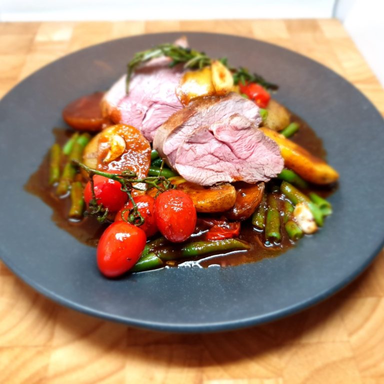 Gericht mit Fleisch, Tomaten, Spargel, Karotten und Kräutern auf schwarzem Teller.