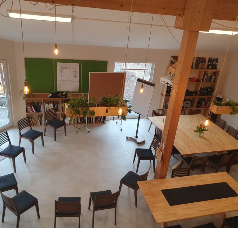 Lichtdurchfluteter Raum mit Tischen, Stühlen und Pflanzen für Workshops.